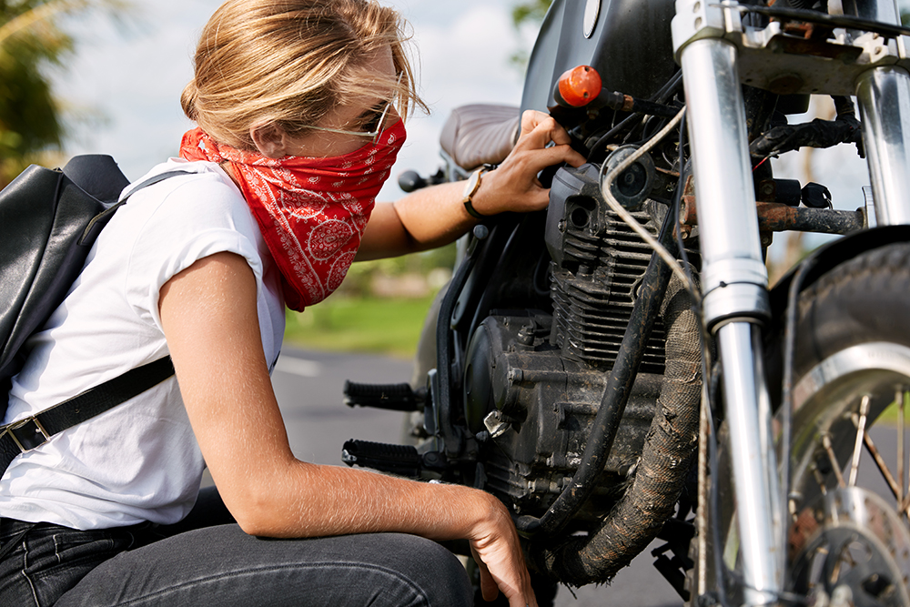 Professional young female biker in casual clothing, wears bandana covedred face, tries to solve mechanical problem with motorbike, stops on countryside road, repairs vehicle. Taking care of bike